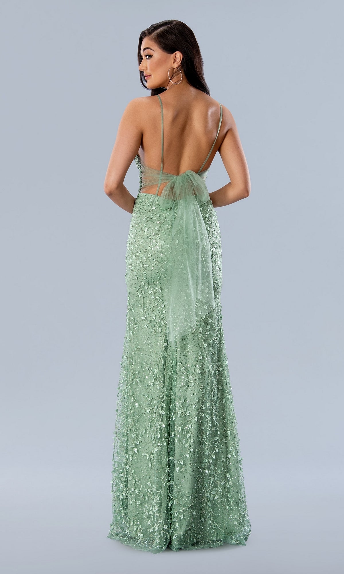 A woman is wearing the Long Prom Dress 24185 by Stella Couture, a light green beaded formal gown with thin straps, a sheer bow, and an open back, standing against a plain light gray background.